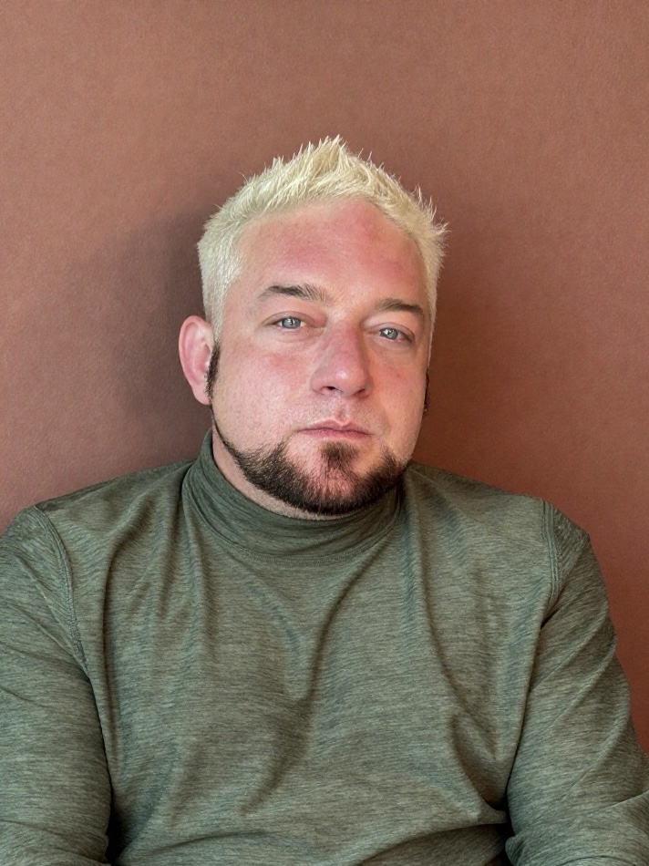 Person with short, bleached hair and a goatee, wearing a green shirt, looking at the camera against a brown background.