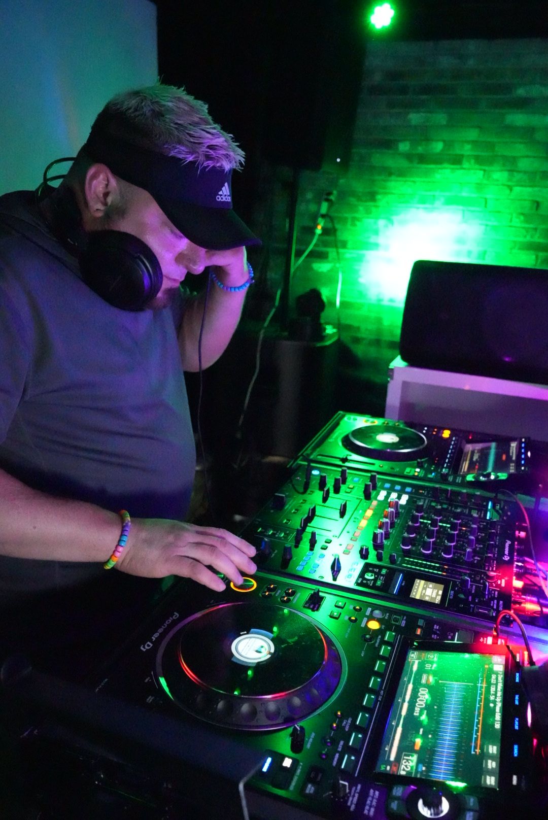 DJ wearing headphones and a cap, operating a mixing console with colorful lights, in a dark club setting.