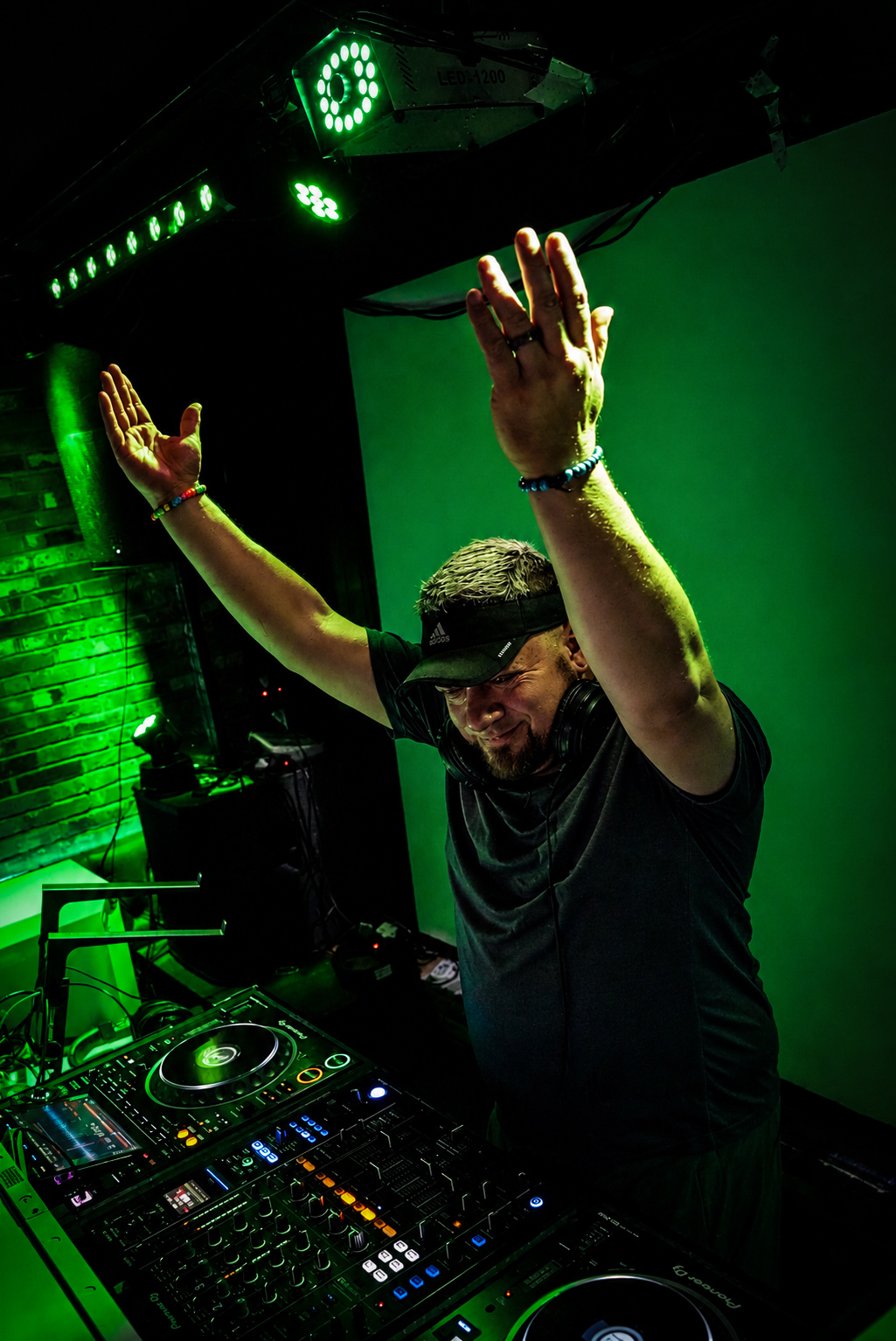 DJ with arms raised, wearing headphones, in a dark club with green lighting and DJ equipment visible.
