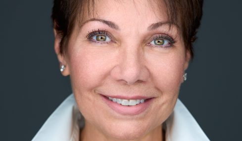 Portrait of a woman with short dark hair, blue eyes, smiling, wearing a white collared shirt and black sweater, against a dark background.