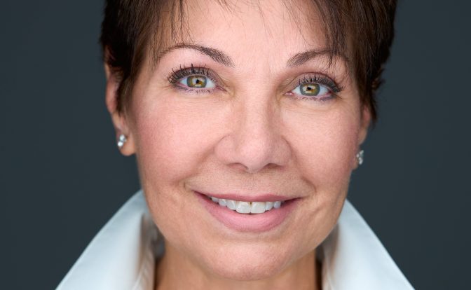 Portrait of a woman with short dark hair, blue eyes, smiling, wearing a white collared shirt and black sweater, against a dark background.