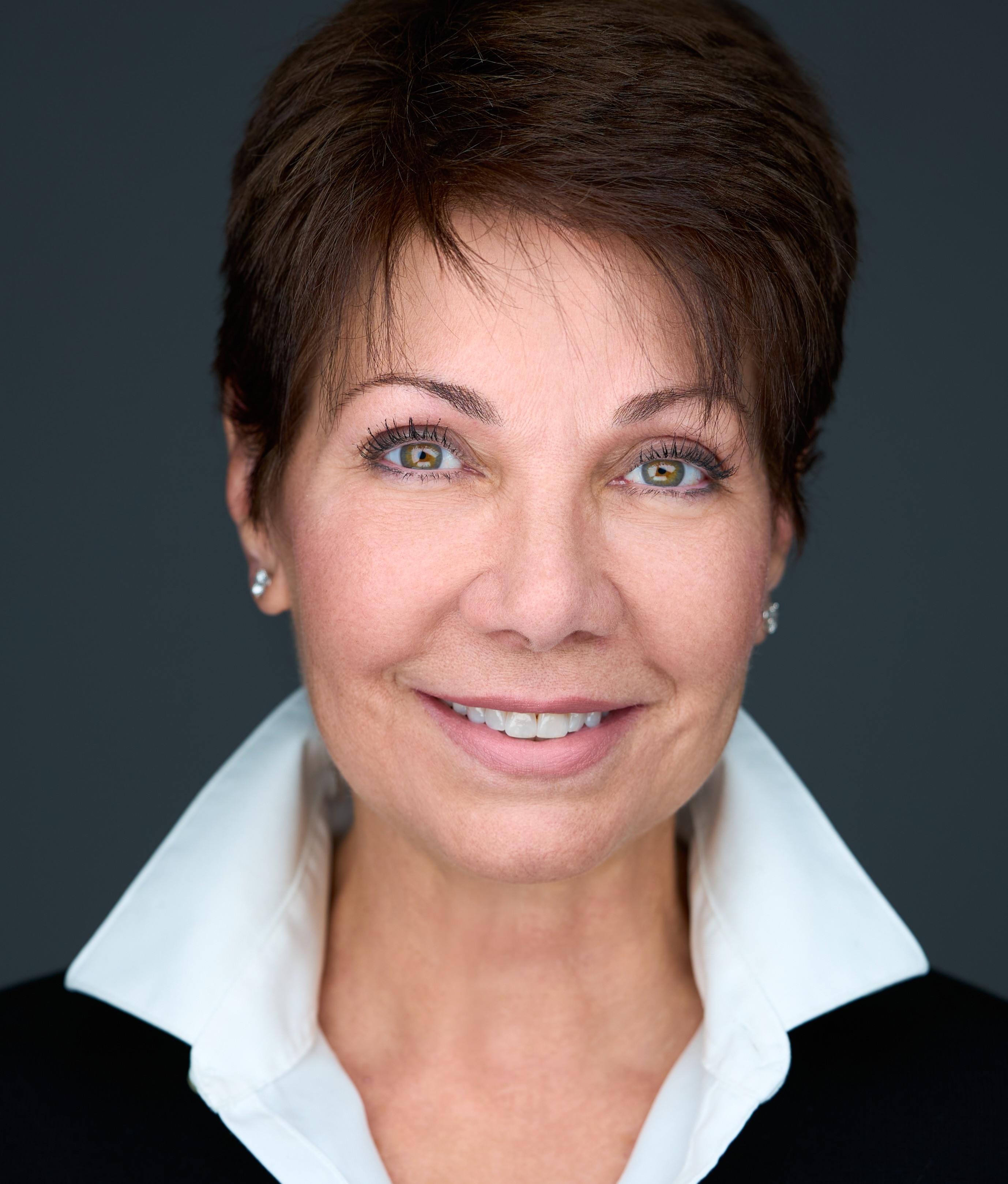Portrait of a woman with short dark hair, blue eyes, smiling, wearing a white collared shirt and black sweater, against a dark background.