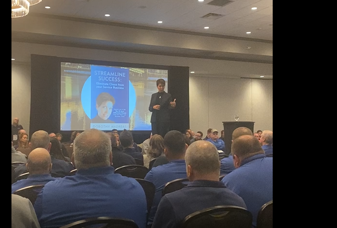 Person standing on stage giving a presentation to an audience in a conference room.