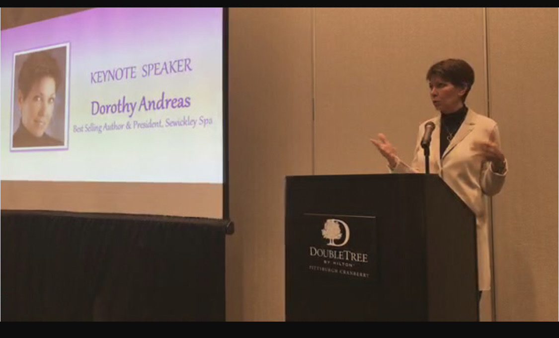 Woman speaking at a podium during a presentation, with a slide showing her photo and name on the screen.