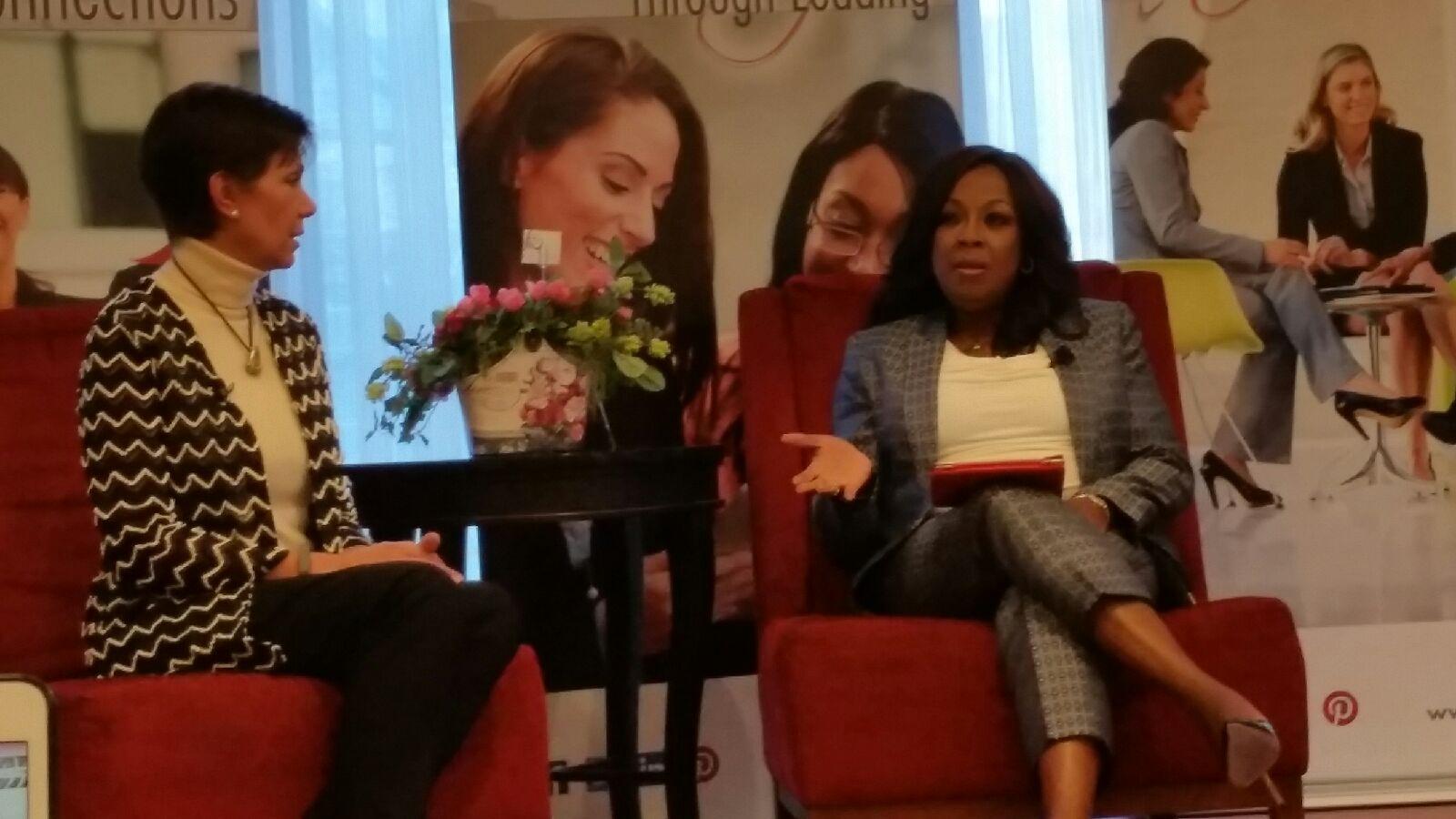 Two women seated on red chairs engaged in conversation, with a small table and a backdrop featuring large images of women.
