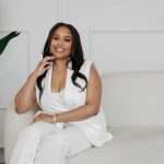 Woman sitting on a sofa, smiling, wearing a white outfit, with a large plant in the background.