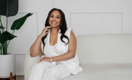 Woman sitting on a sofa, smiling, wearing a white outfit, with a large plant in the background.