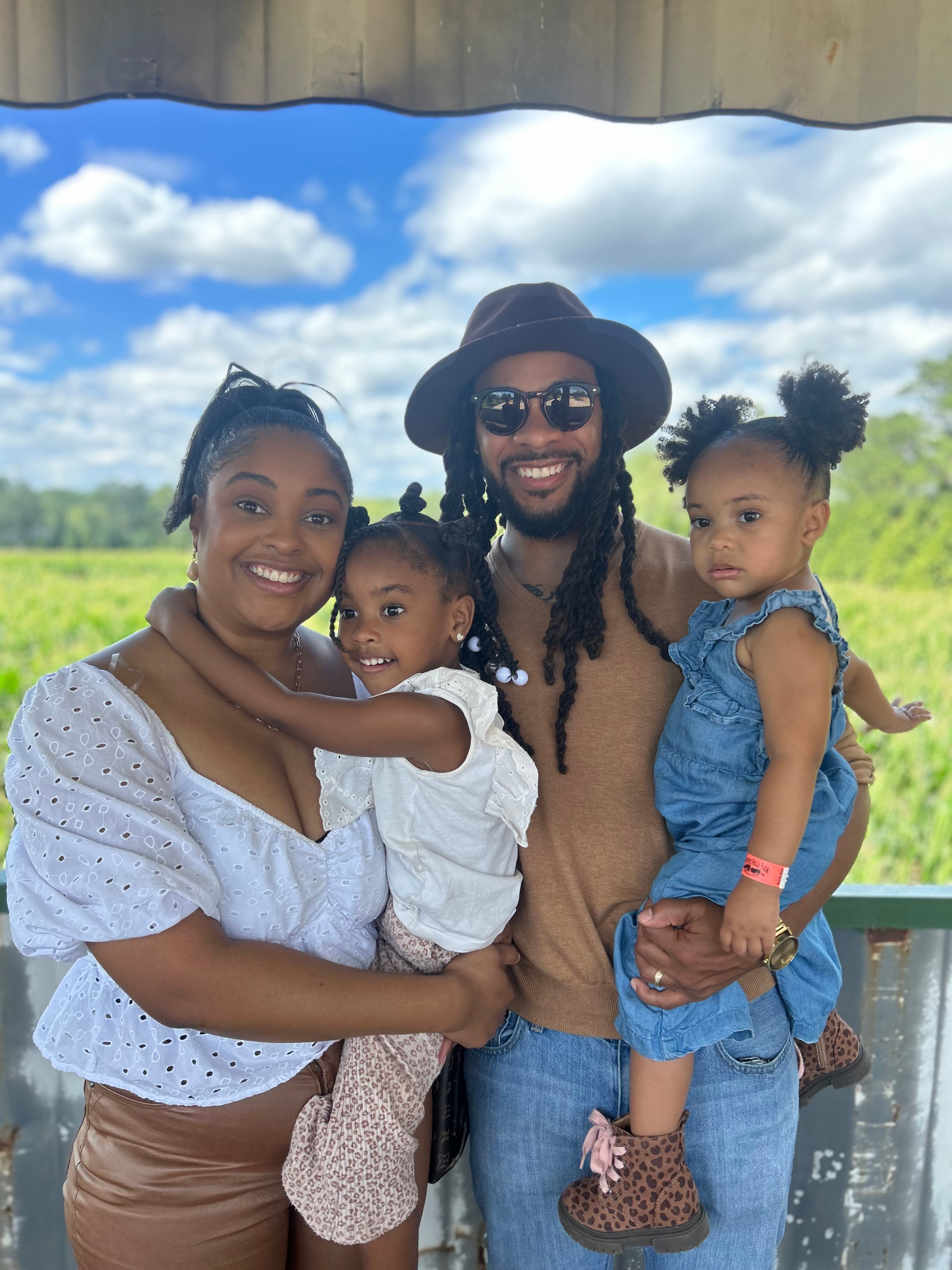 Family of four outdoors, smiling, with blue sky and clouds, woman holding a girl, man holding another girl, all smiling.
