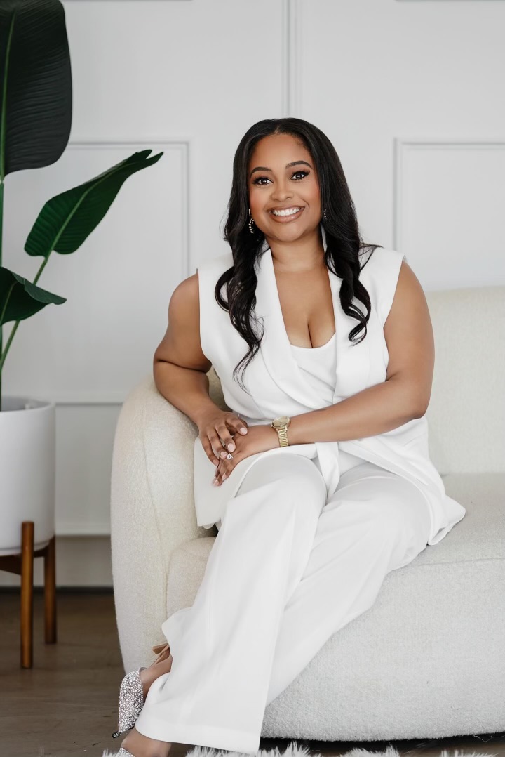 Woman sitting on a white sofa, smiling, wearing white outfit, in a modern room with a large plant.