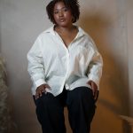 Woman with natural hair wearing a white oversized shirt and black pants, sitting against a plain wall.