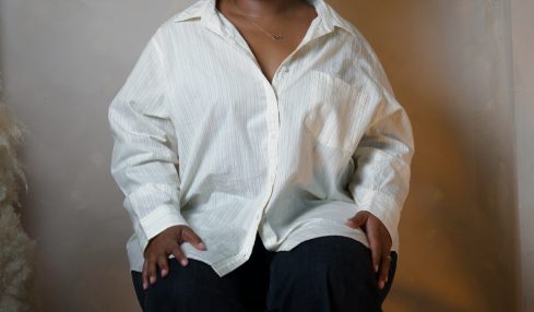 Woman with natural hair wearing a white oversized shirt and black pants, sitting against a plain wall.