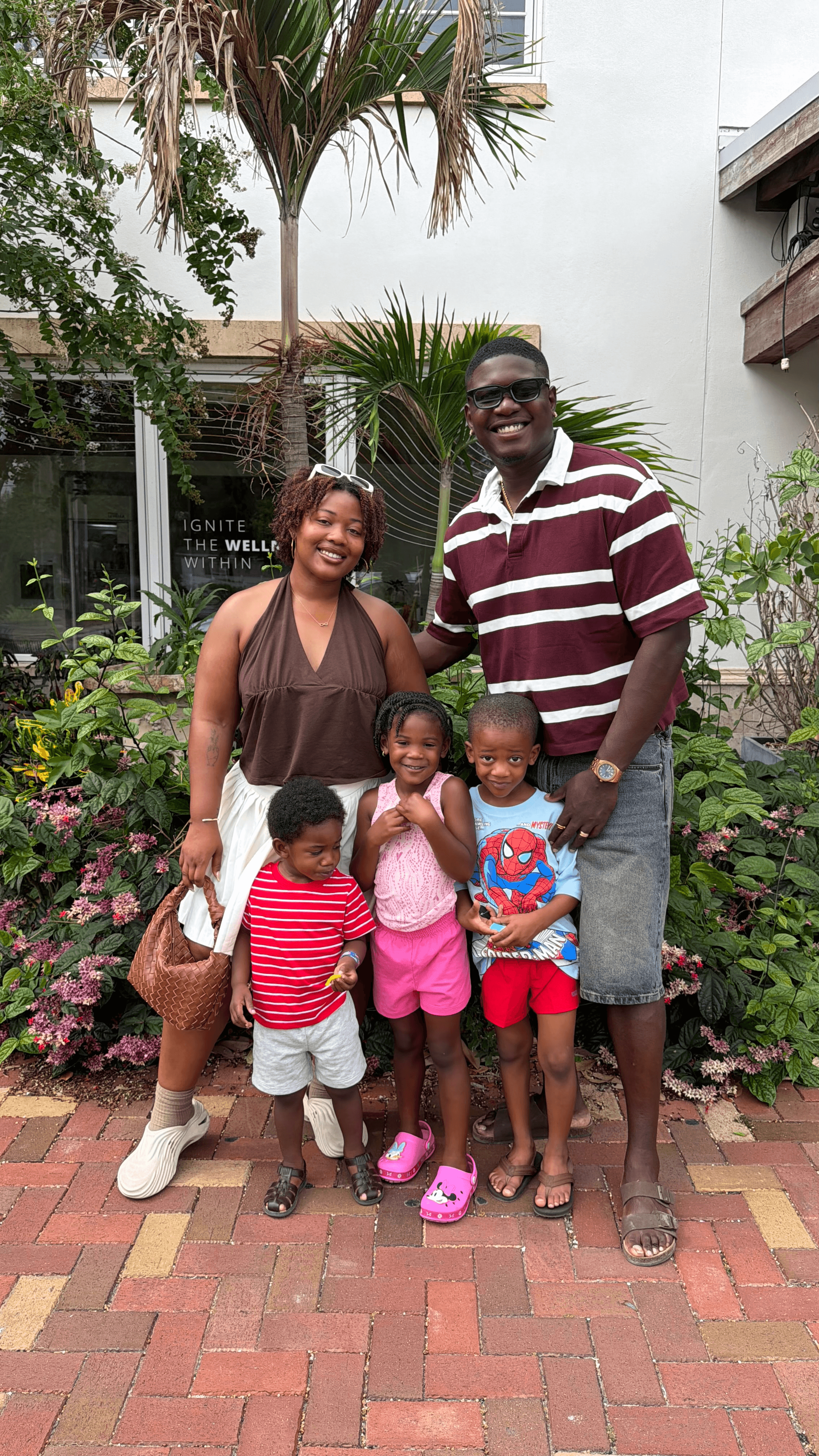 Group of five people, two adults and three children, standing outdoors on a brick path with plants and trees behind them.