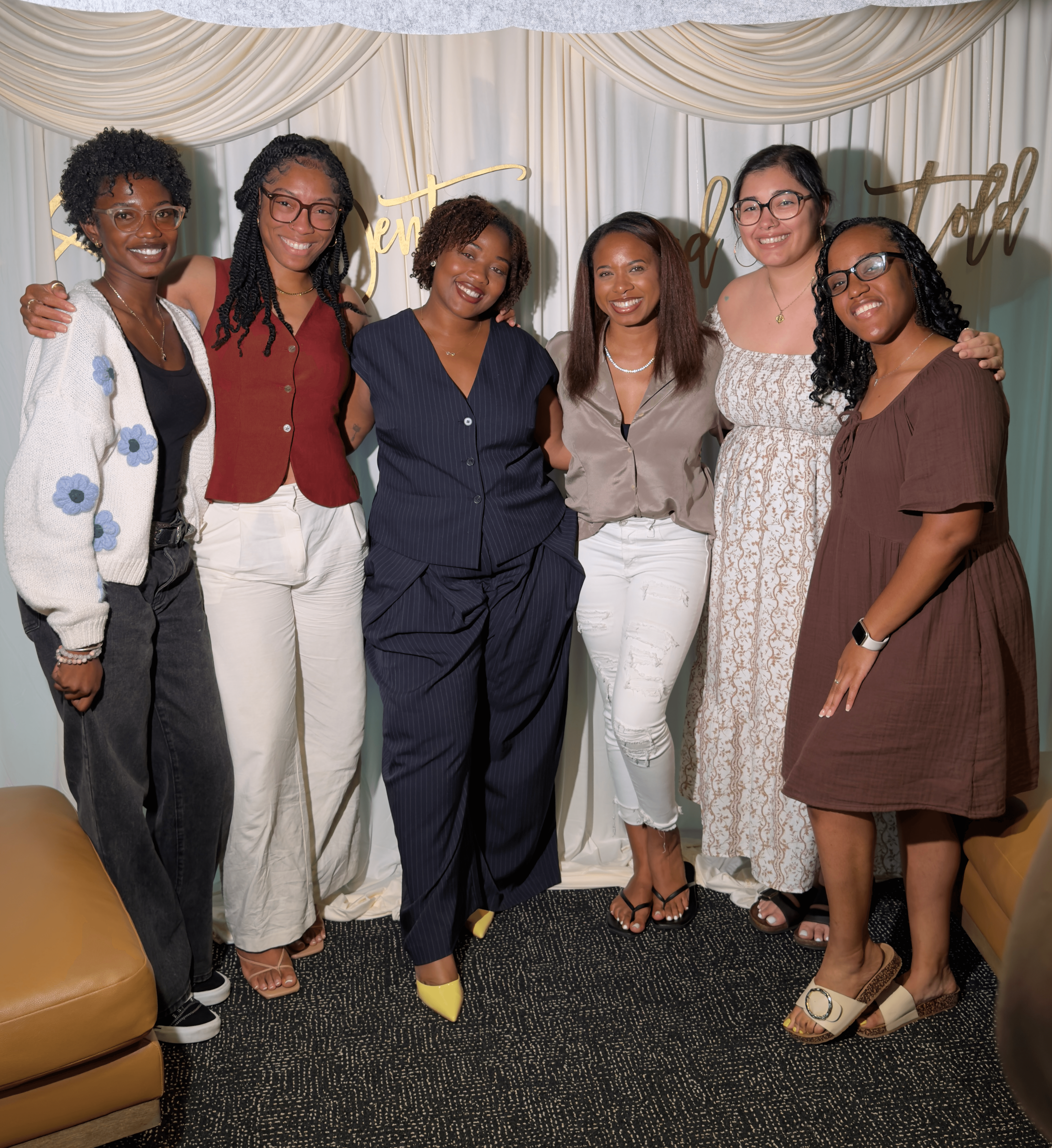 Six women standing together indoors, smiling, with a decorated backdrop behind them.