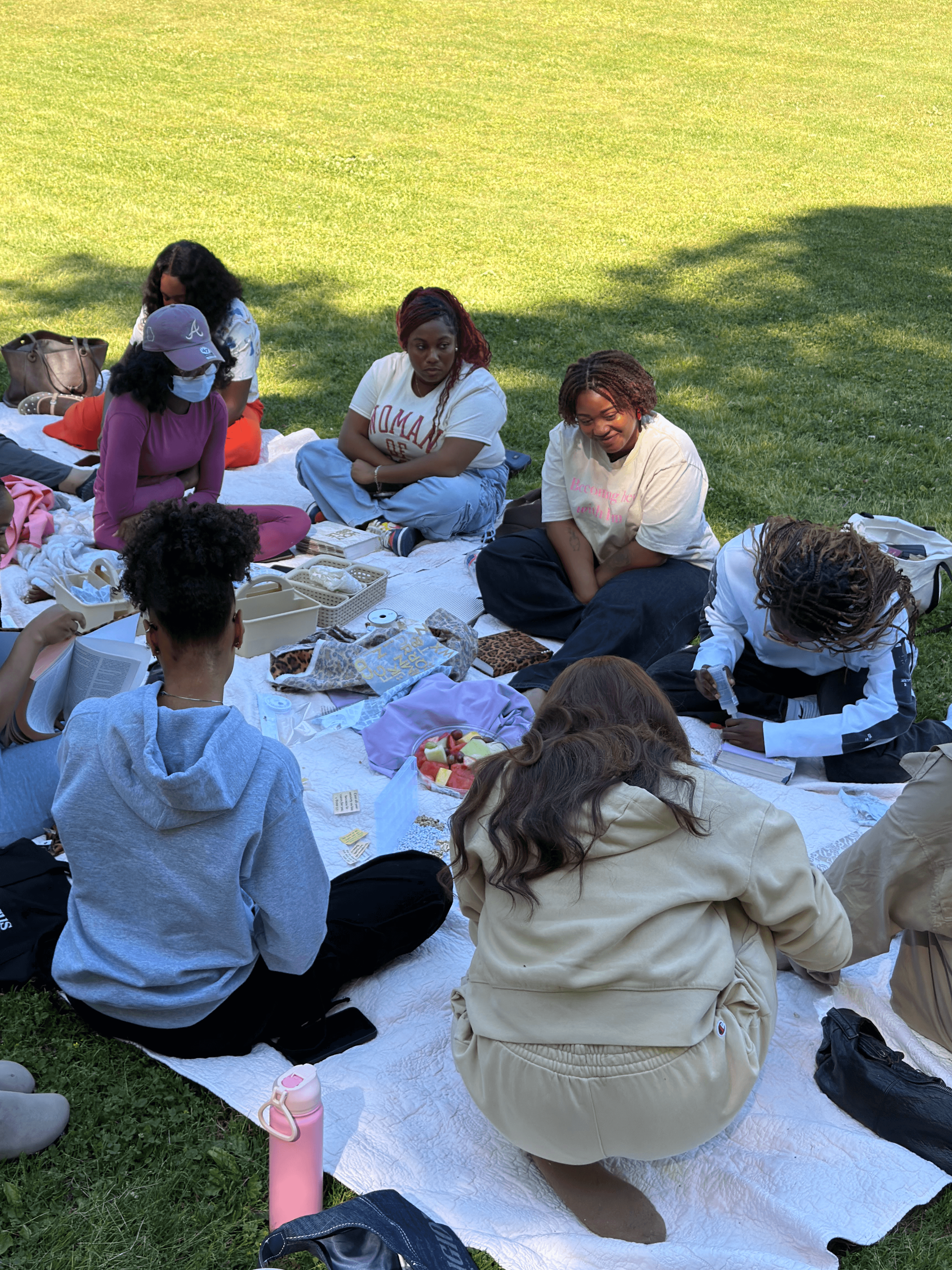 Group of people sitting on a white blanket outdoors on grass, engaging in a discussion or activity, with various items around them.