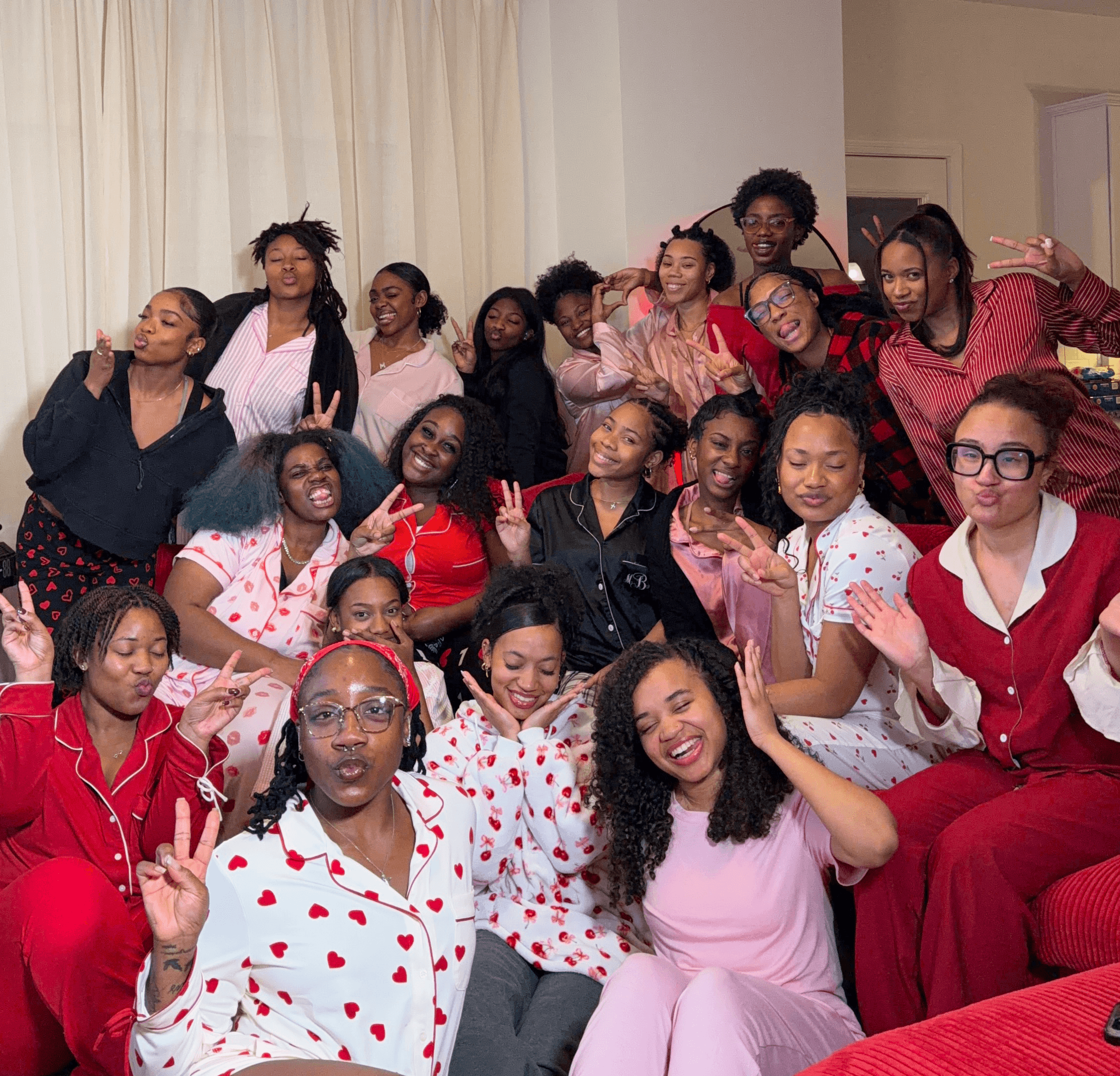 Group of women and girls smiling and posing indoors, some making peace signs, in casual and pajamas, in a living room.