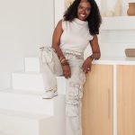 Woman with curly hair in white sleeveless top and ripped jeans, smiling, leaning on a cabinet in a bright room.