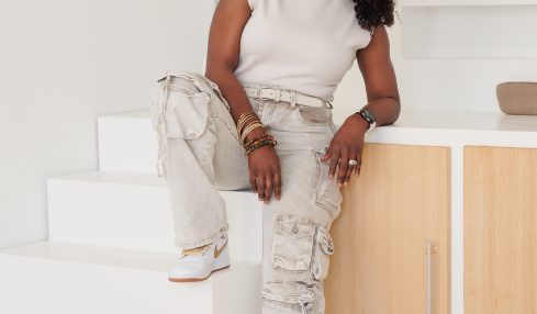 Woman with curly hair in white sleeveless top and ripped jeans, smiling, leaning on a cabinet in a bright room.