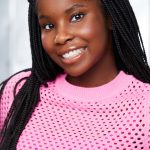 Young woman with braided hair smiling, wearing a pink perforated top, indoors with a blurred background.