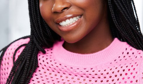Young woman with braided hair smiling, wearing a pink perforated top, indoors with a blurred background.