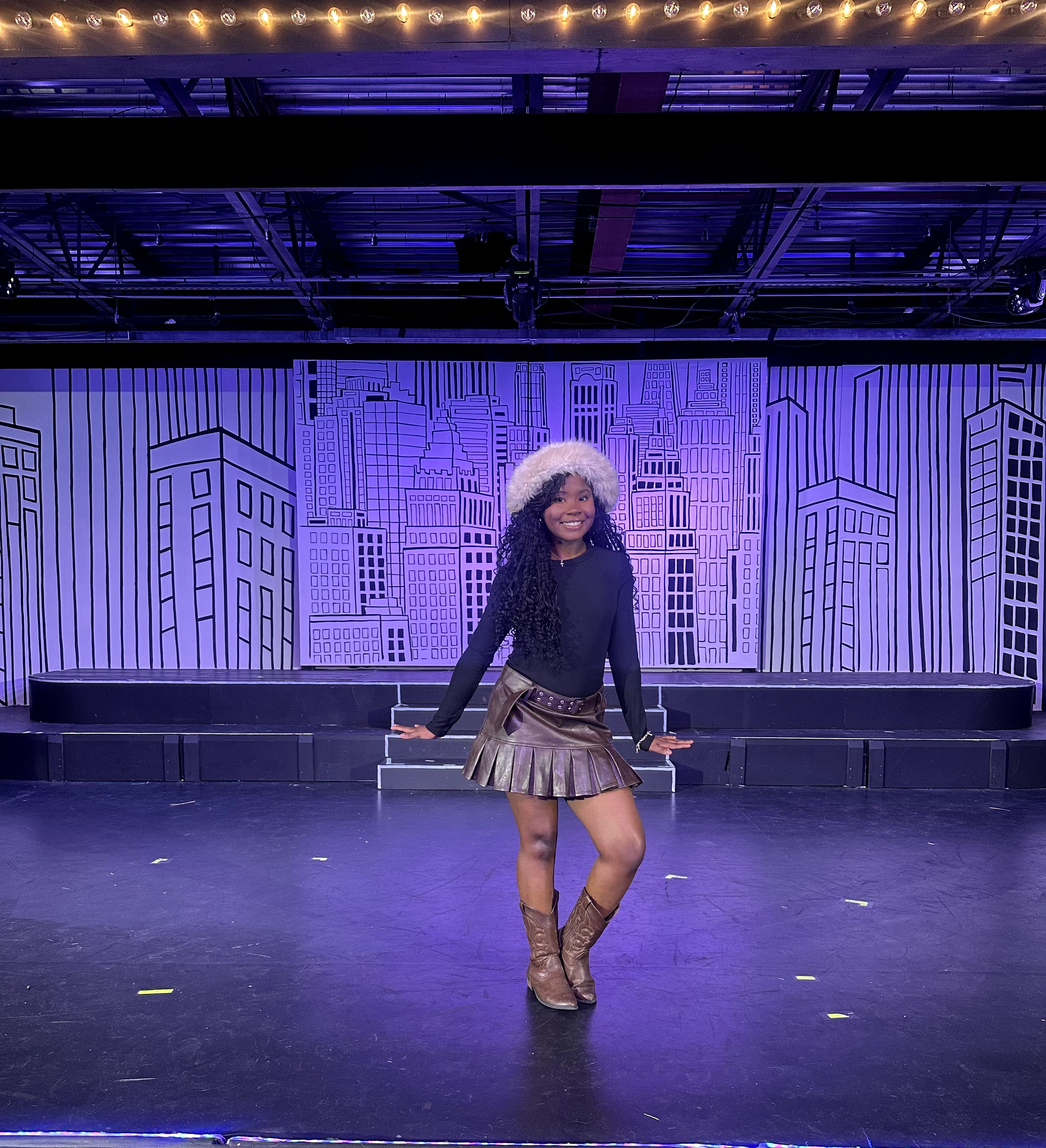 Person standing on stage with cityscape backdrop, wearing a hat, sweater, skirt, and boots, smiling, with stage lights overhead.