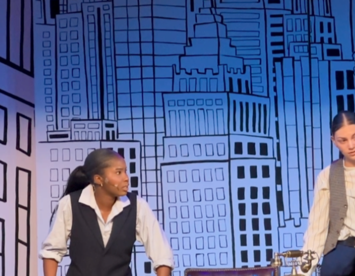 Two women on stage with a cityscape backdrop, one looking to the side, the other partially visible, stage props visible.