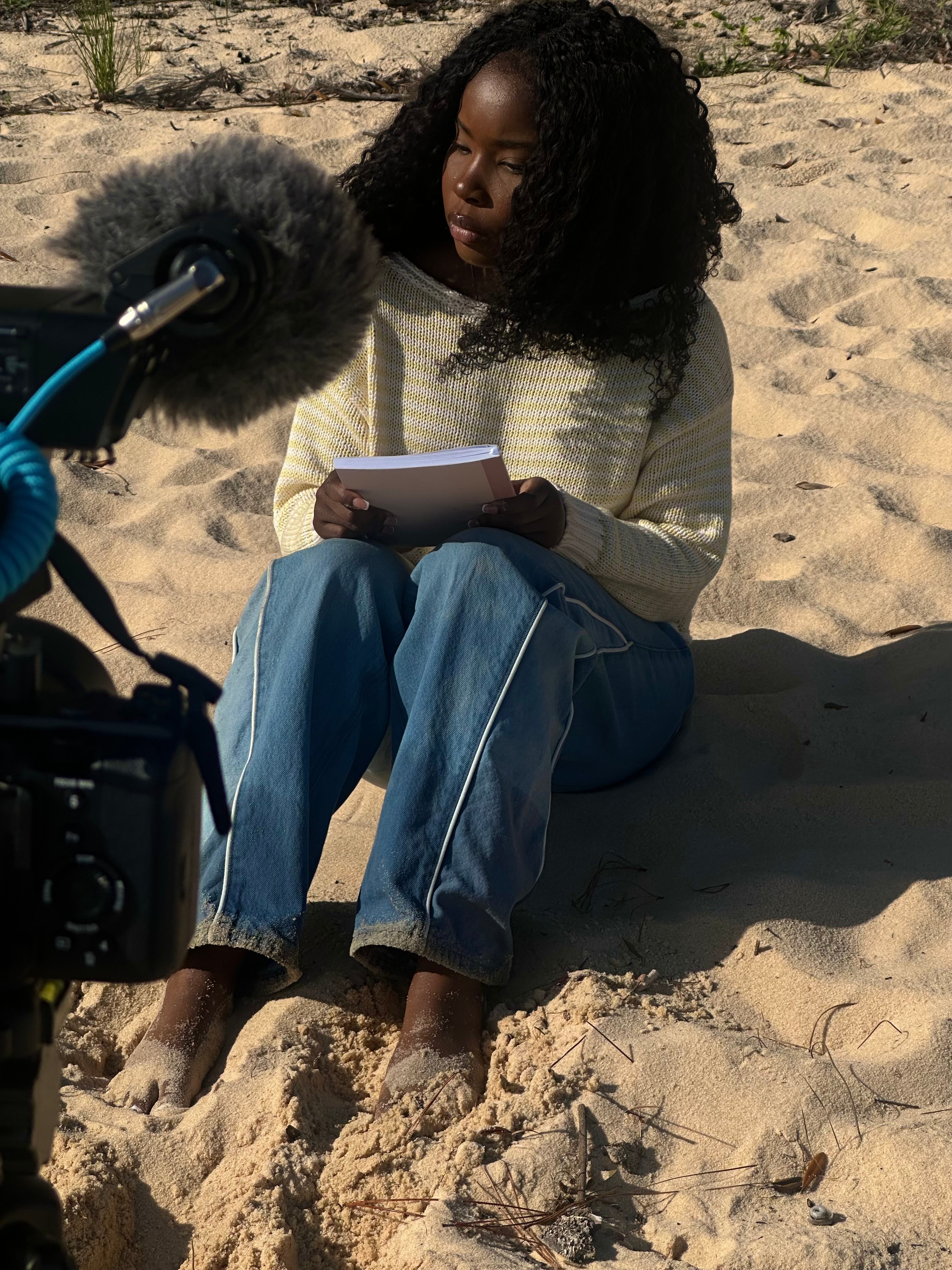 Young woman with curly hair sitting on sand, reading a notebook, with a camera in the foreground.