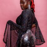 Woman with red braids wearing a black dress with colorful dots and sequined skirt, posing against pink background.