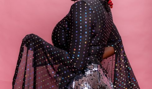 Woman with red braids wearing a black dress with colorful dots and sequined skirt, posing against pink background.