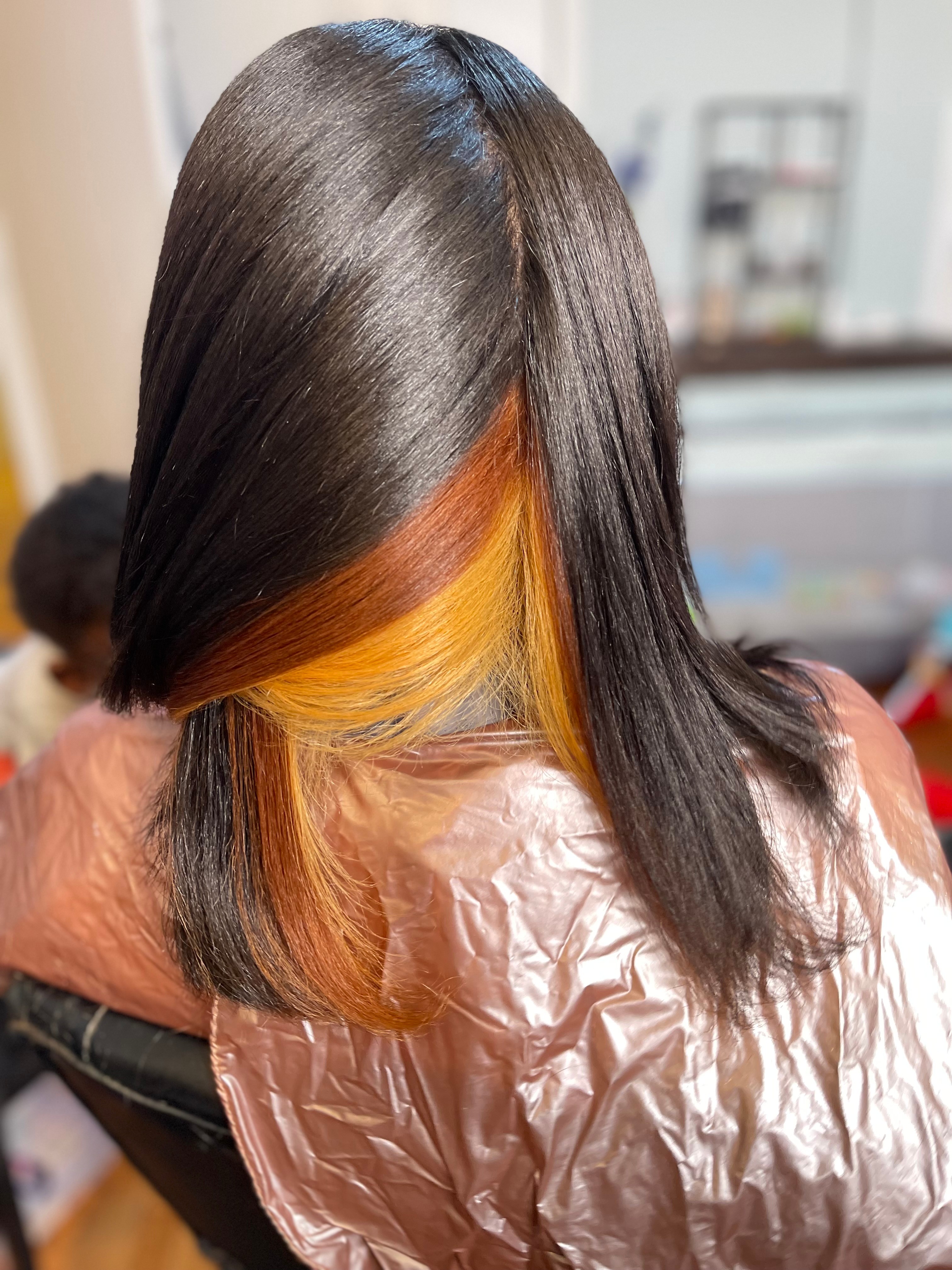 Person with straight, dark hair and orange highlights, sitting in a salon chair, with a cape around shoulders.