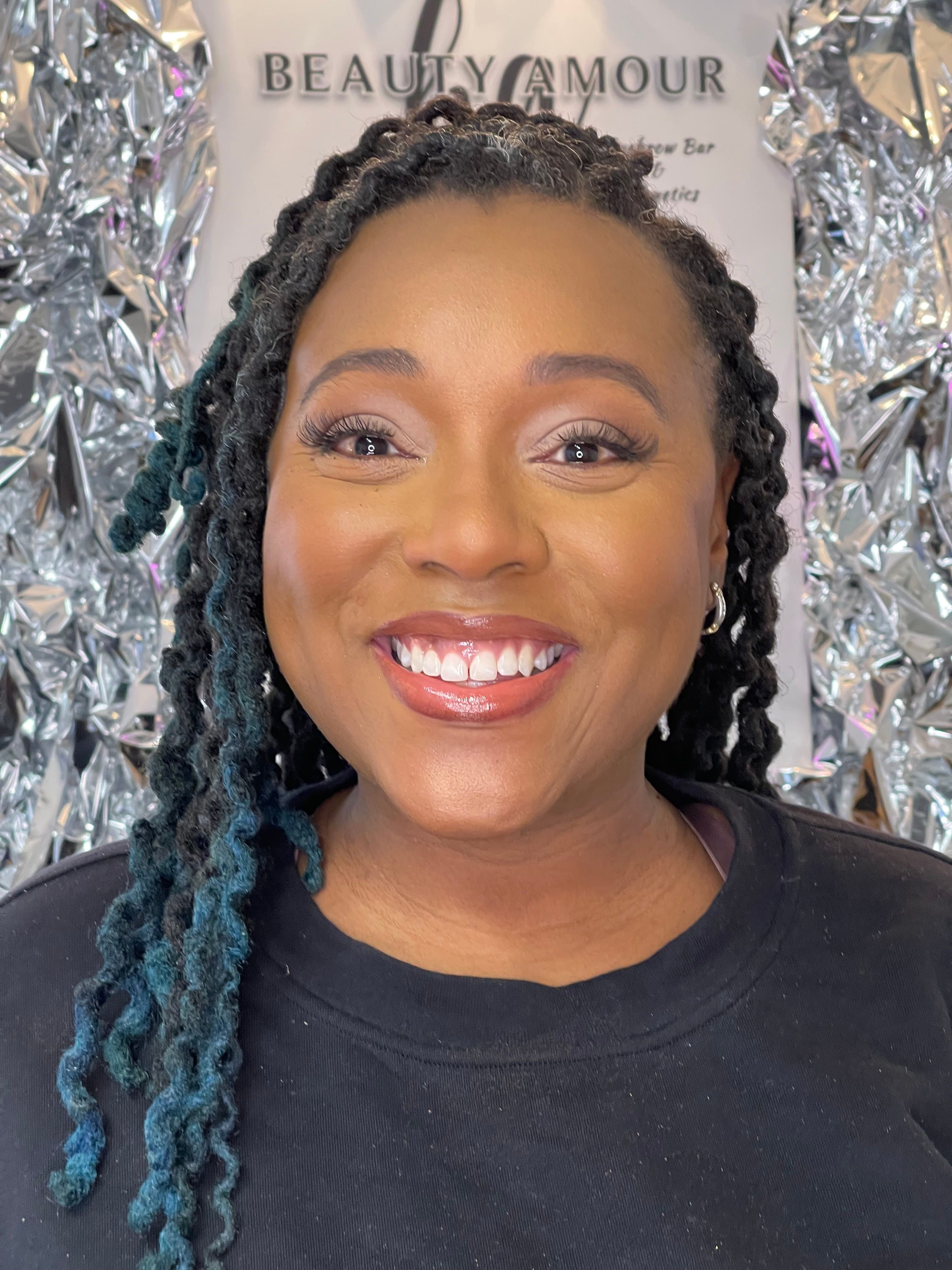 Smiling woman with braided hair, wearing a black shirt, in front of a silver foil background.