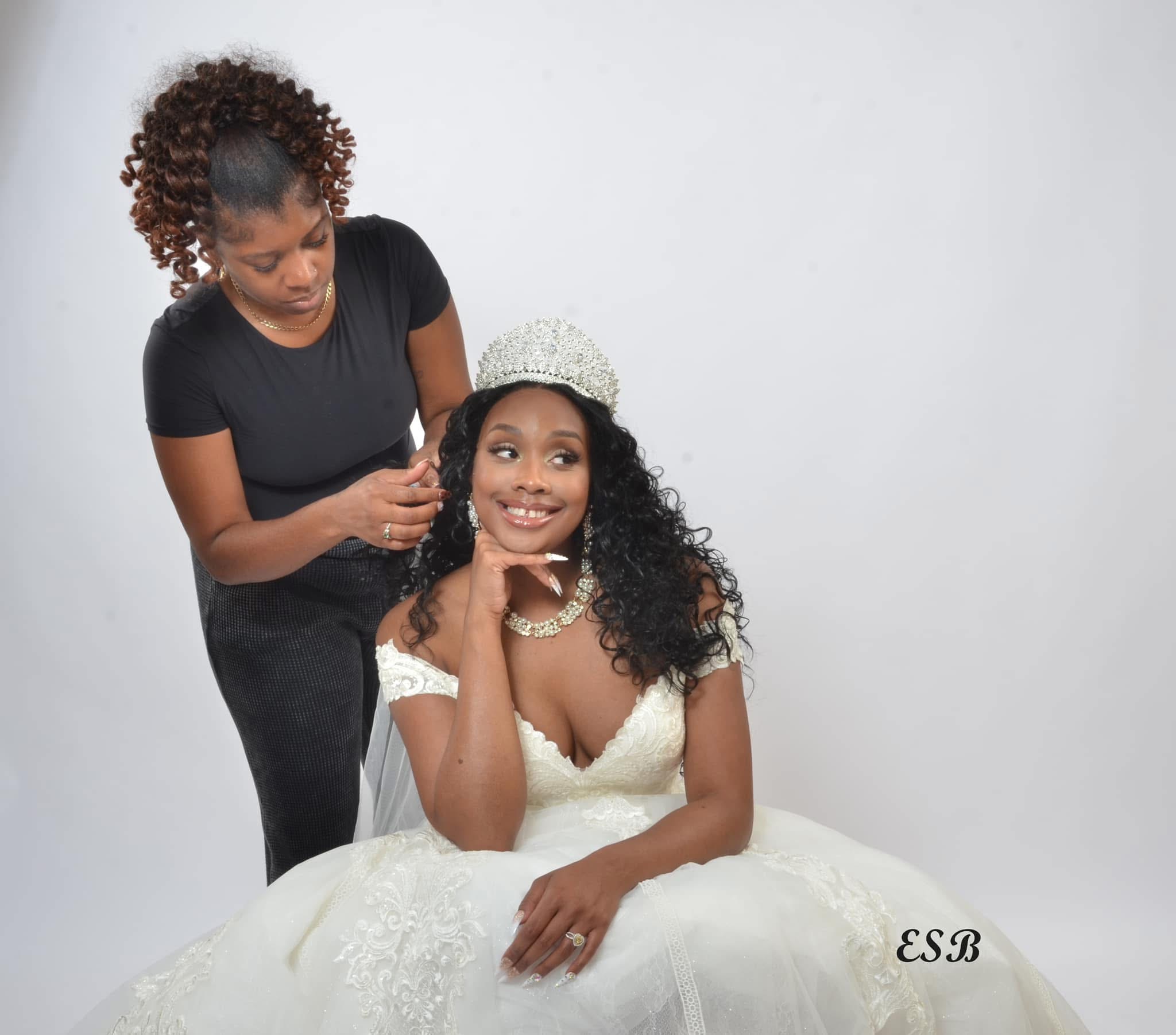 A woman in a wedding dress sits smiling, while another woman styles her hair against a plain white background.