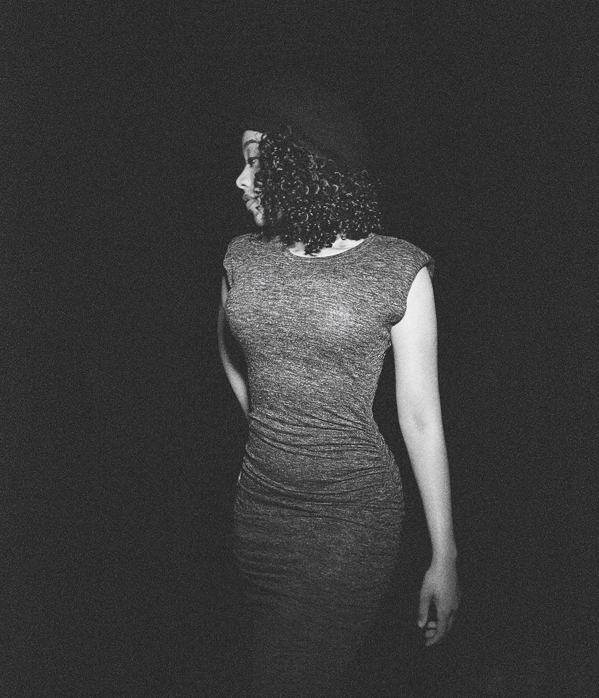 Woman with curly hair in a sleeveless dress, standing sideways against a dark background.
