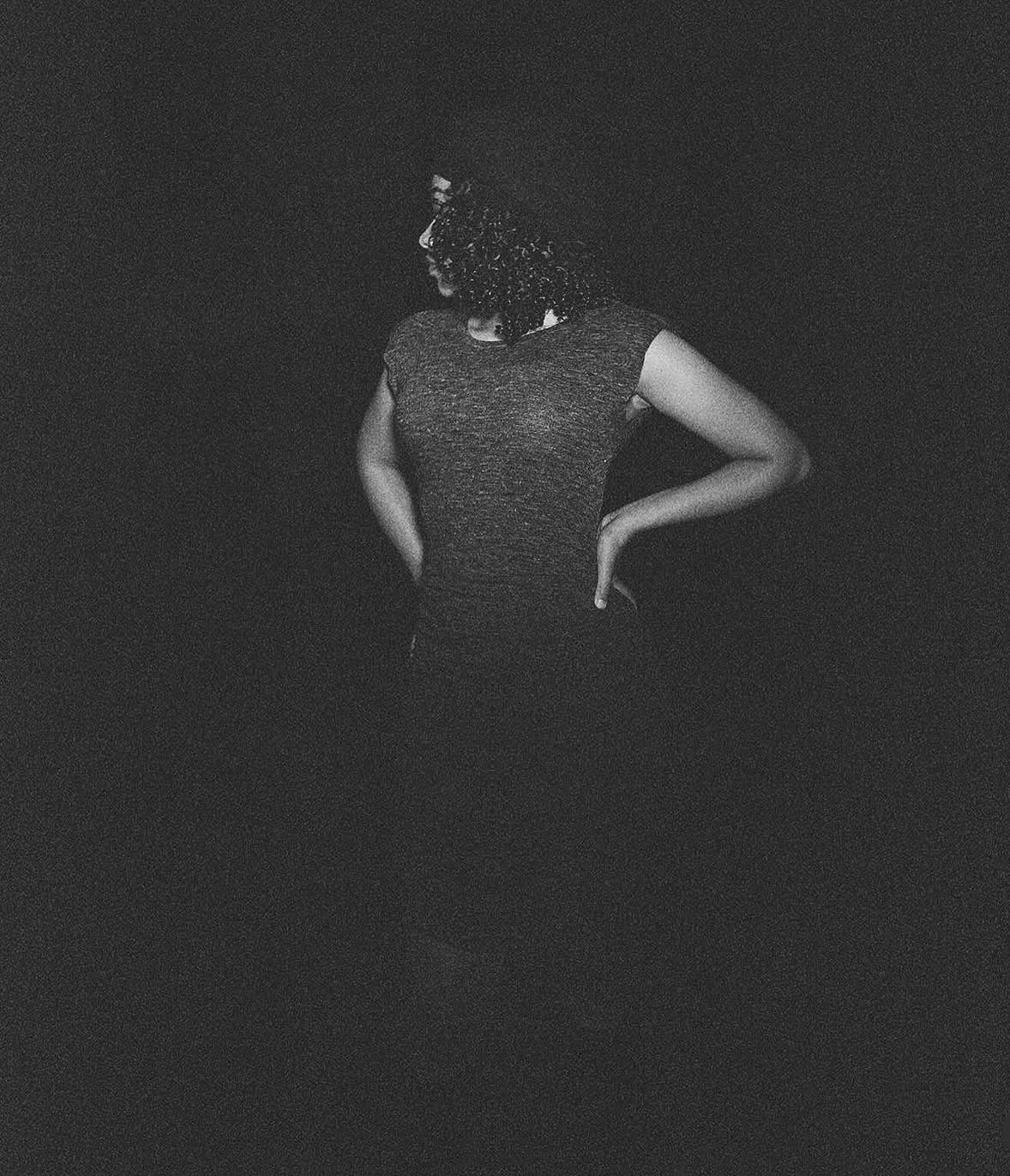 Person with curly hair facing away, hands on hips, wearing a sleeveless dress, in a dark background.
