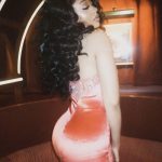 Woman with curly hair in a pink dress, turned sideways, in a dimly lit room with warm lighting.
