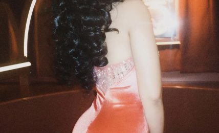 Woman with curly hair in a pink dress, turned sideways, in a dimly lit room with warm lighting.