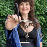 Woman with curly hair holding a sword, smiling, outdoors with green foliage background.