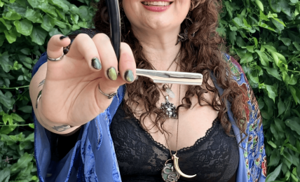 Woman with curly hair holding a sword, smiling, outdoors with green foliage background.