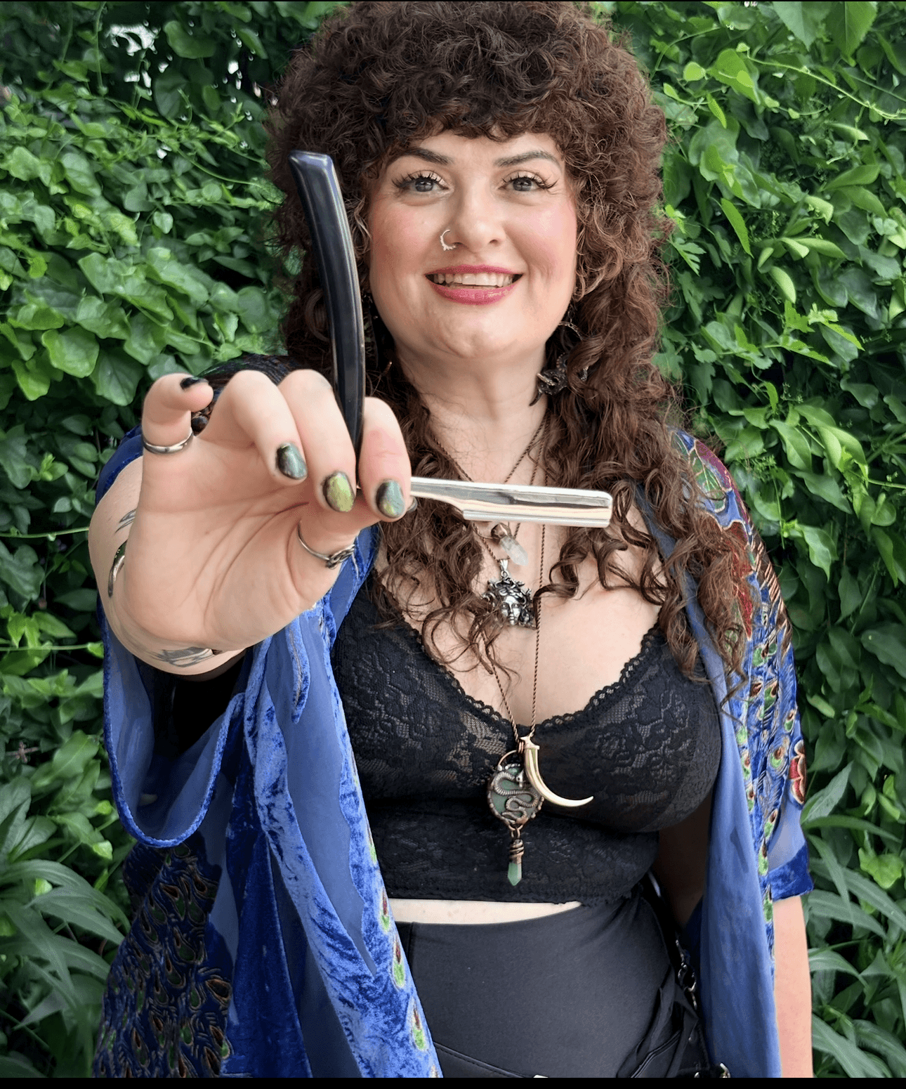 Woman with curly hair holding a sword, smiling, outdoors with green foliage background.
