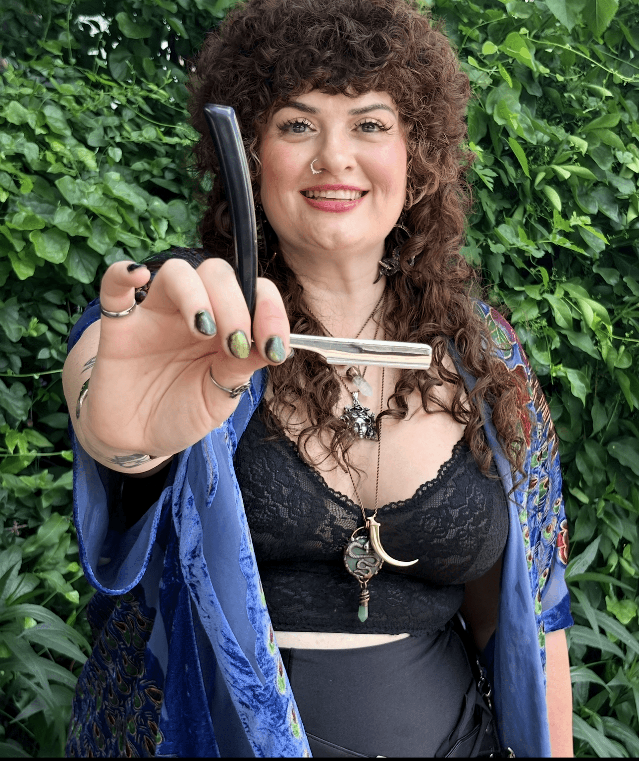 Woman with curly hair holding a sword, smiling, outdoors with green foliage background.