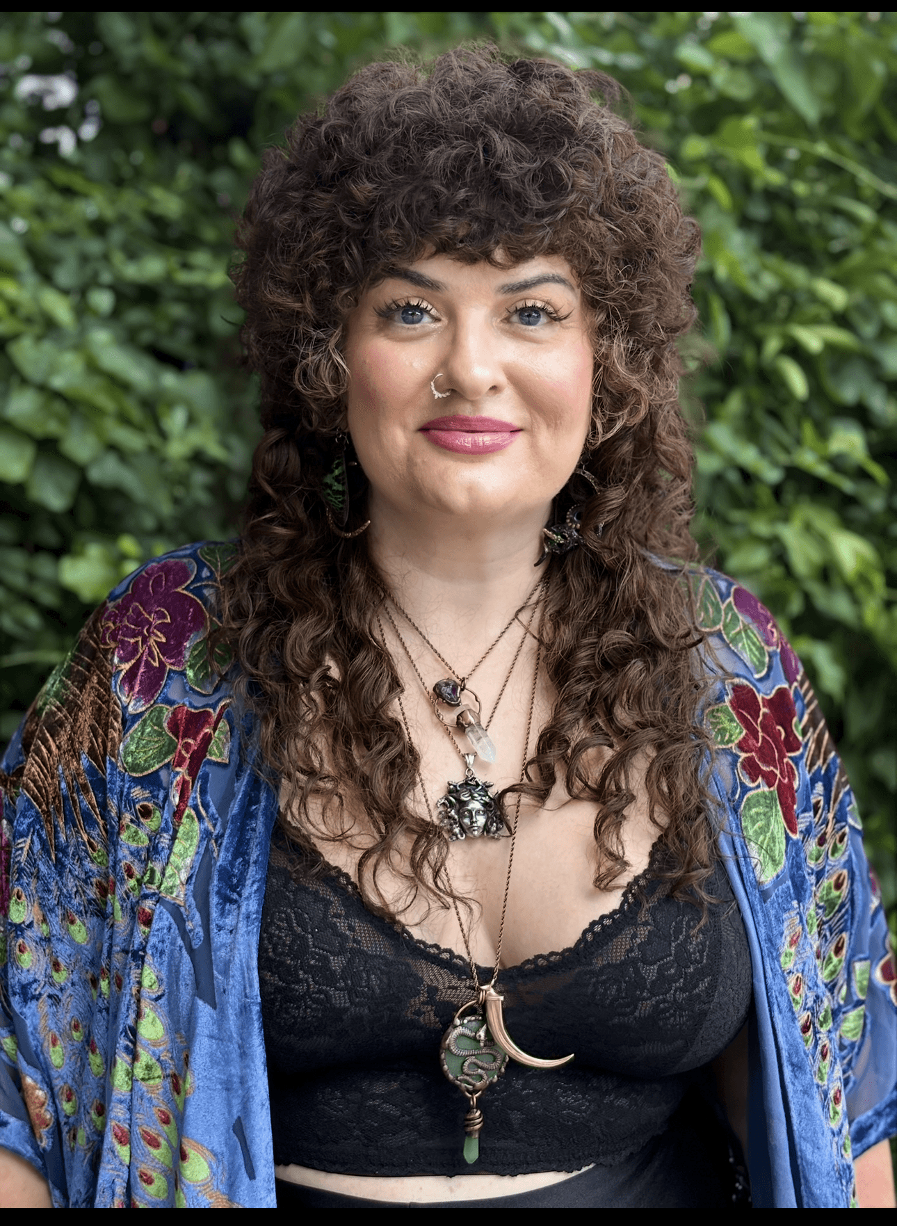 Woman with curly hair wearing a colorful shawl and black top, standing outdoors with greenery in background.