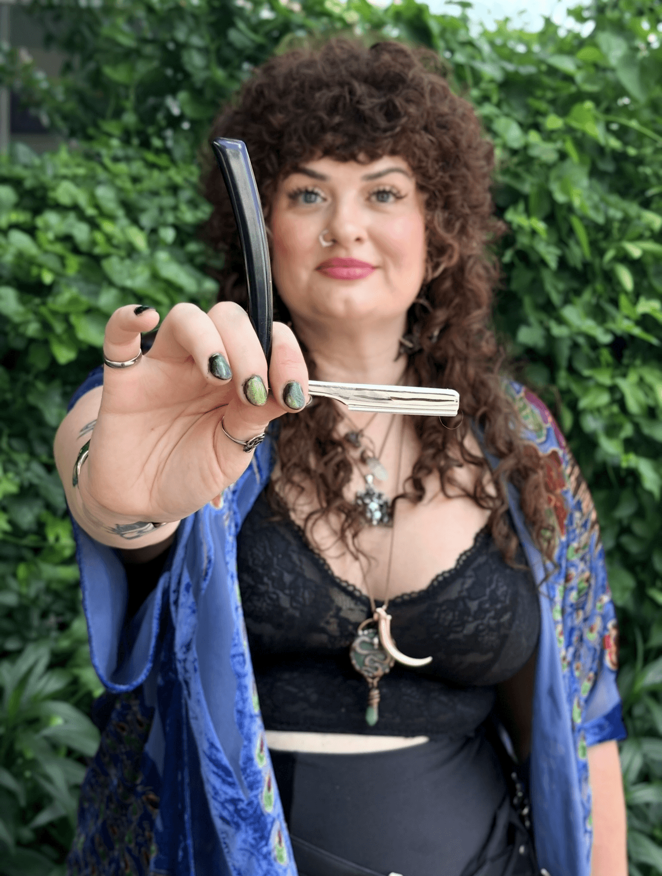 Woman with curly hair holding a knife and a phone, standing outdoors with green foliage behind her.