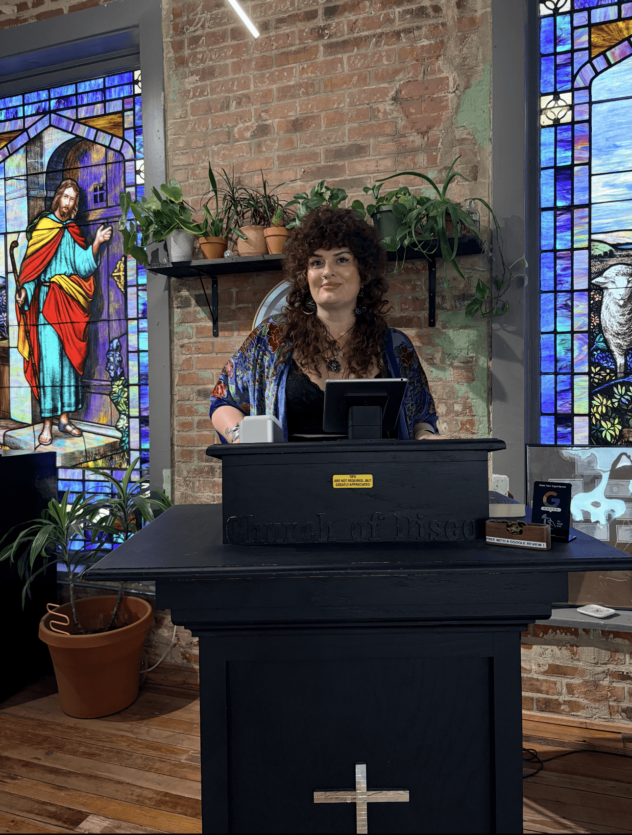 Woman standing behind a pulpit with a cross, in front of stained glass windows, brick wall, and plants.