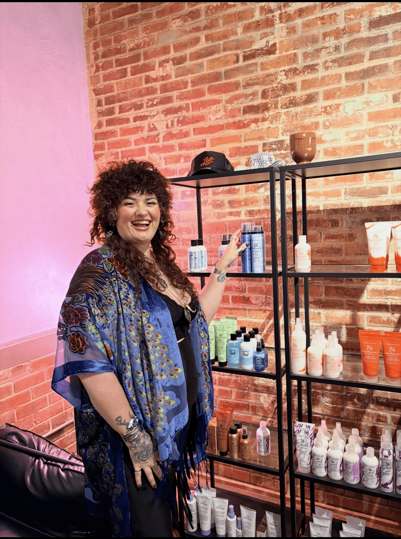 Woman with curly hair smiling, standing in front of shelves with various bottles and containers, brick wall background.