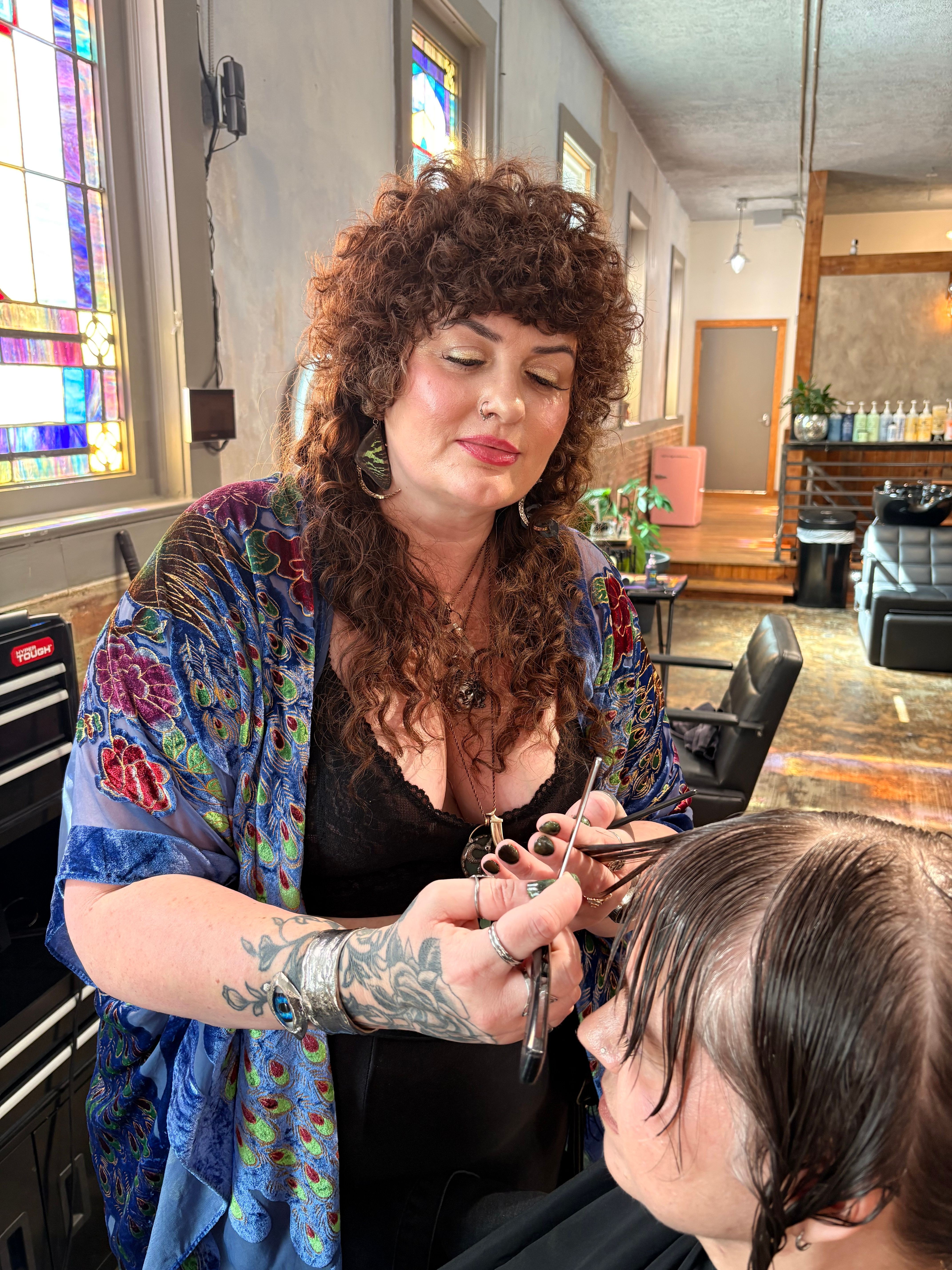 Two women sit at a table in a cafe, one with curly hair and colorful clothing, the other with dark hair, glasses, and tattoos, engaged in conversation.