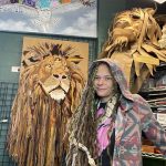 Person with long braided hair and a snake around their shoulders standing next to a lion sculpture made of wood or similar material.