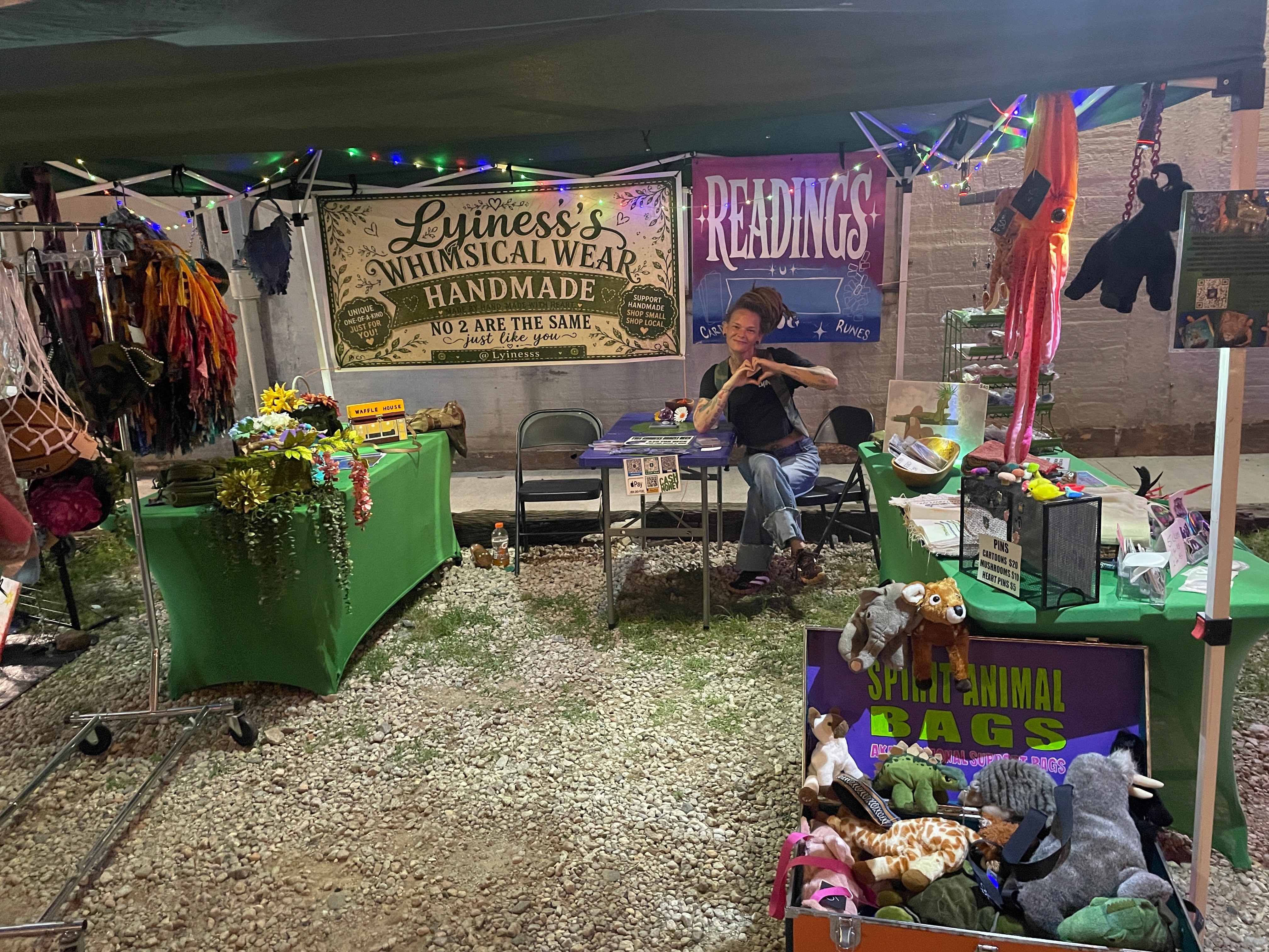 Market stall with handmade items, signs for readings and animal bags, and a person sitting at a table outdoors.