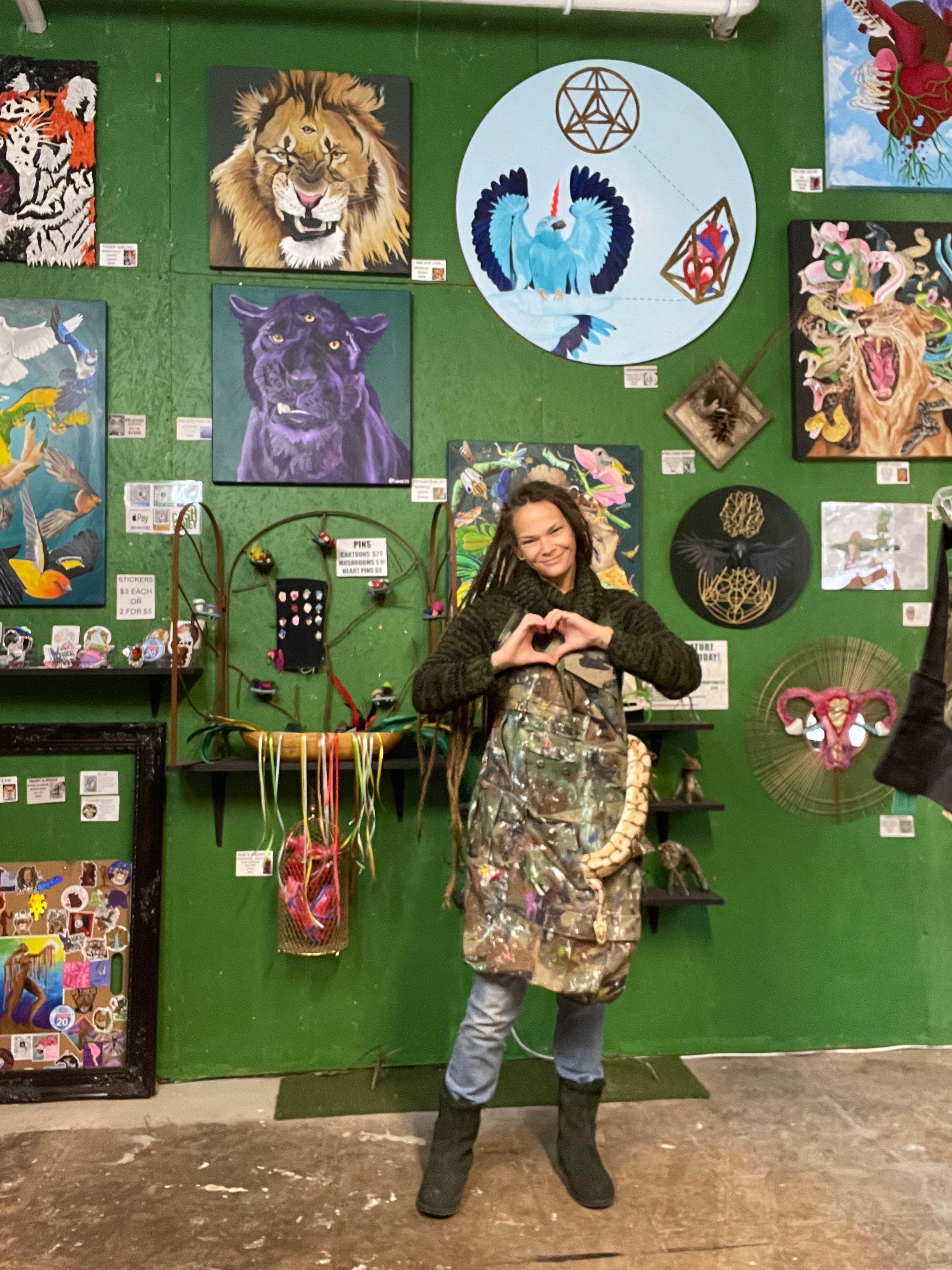 Woman in camouflage coat and jeans standing in art gallery with colorful paintings and sculptures, green wall background.