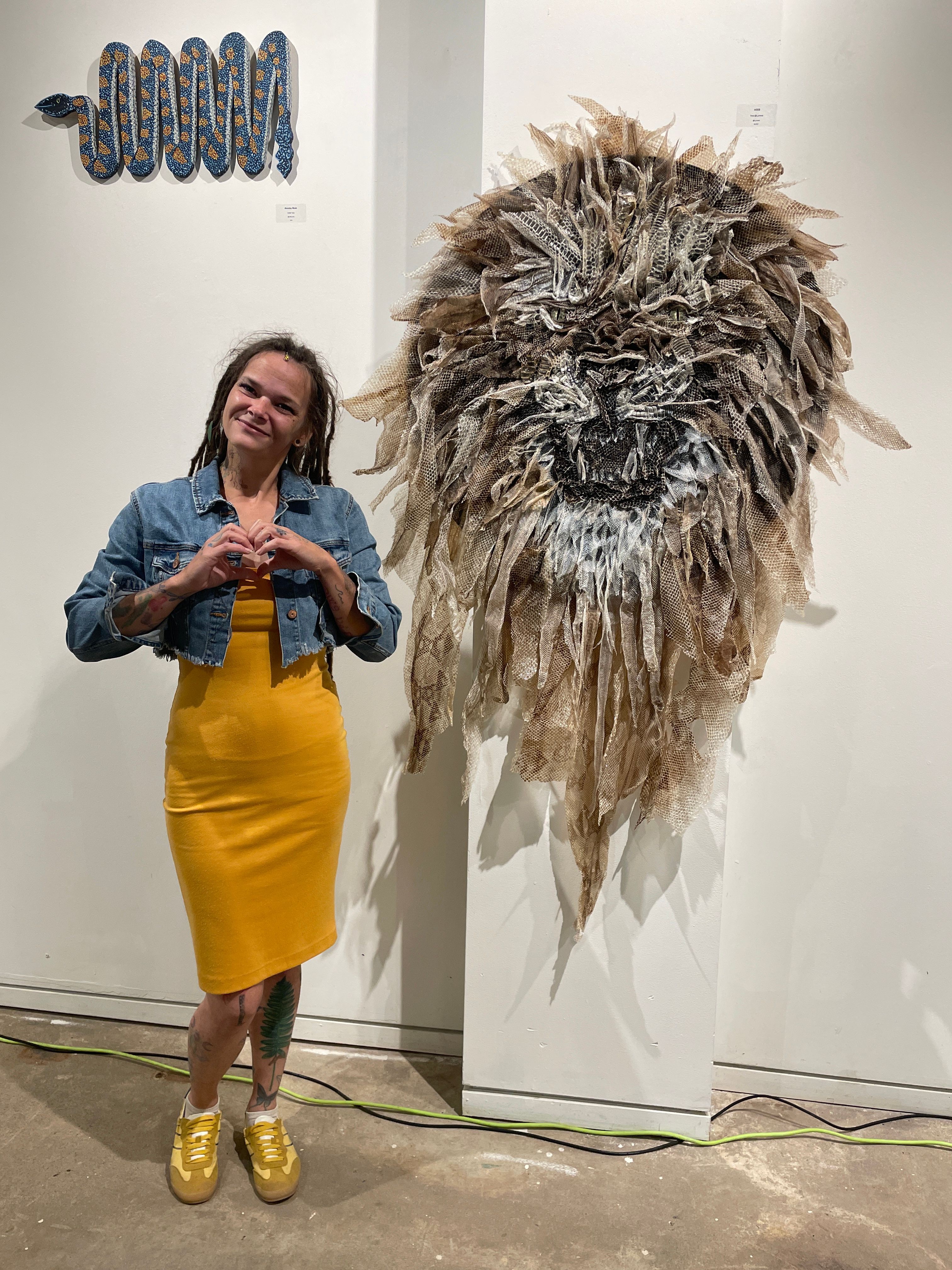 Woman in yellow dress and denim jacket taking a selfie in front of a large feathered art piece on a white wall.