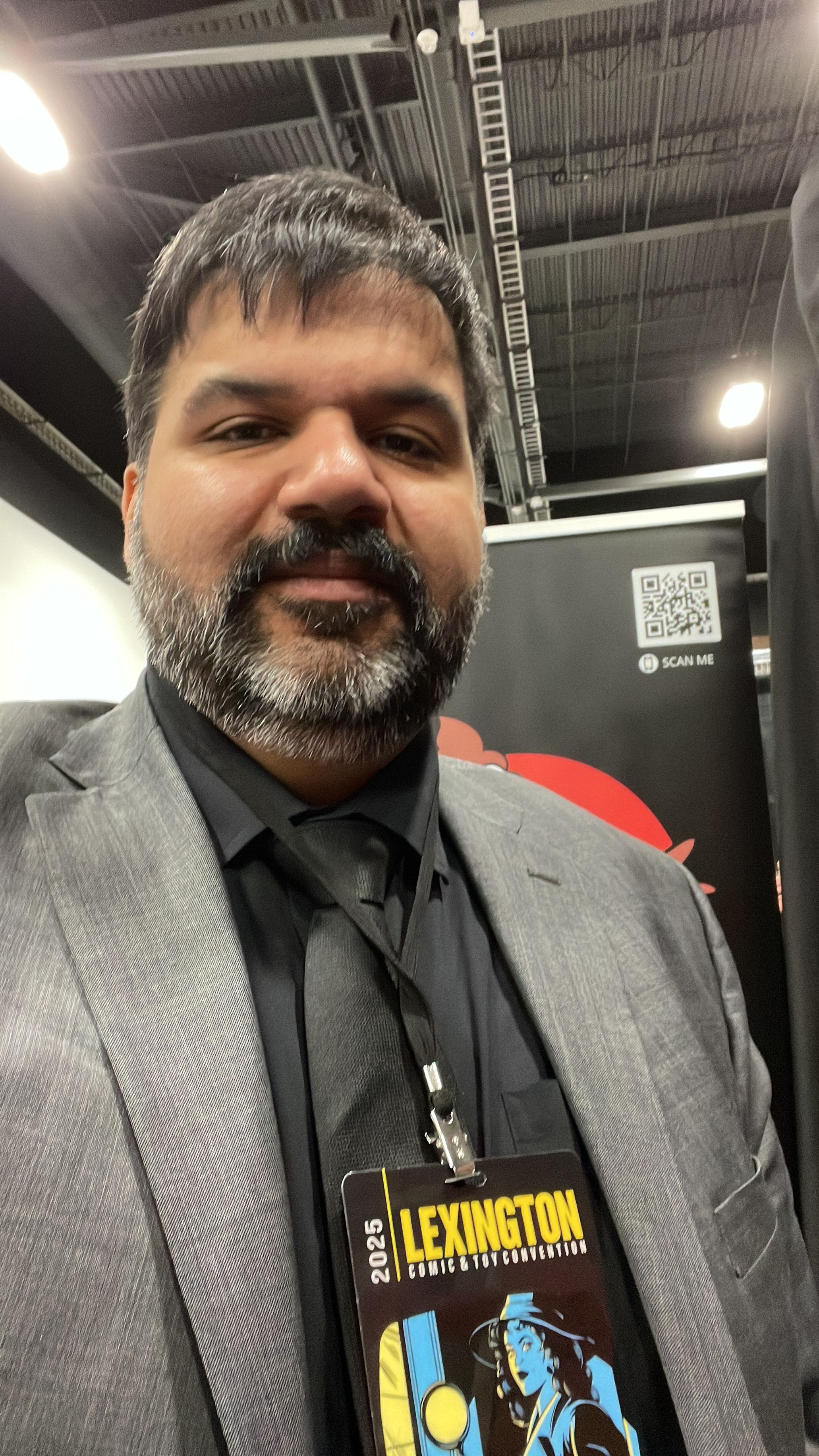 Man with gray hair and beard wearing a gray suit and black shirt, standing indoors with a dark background.