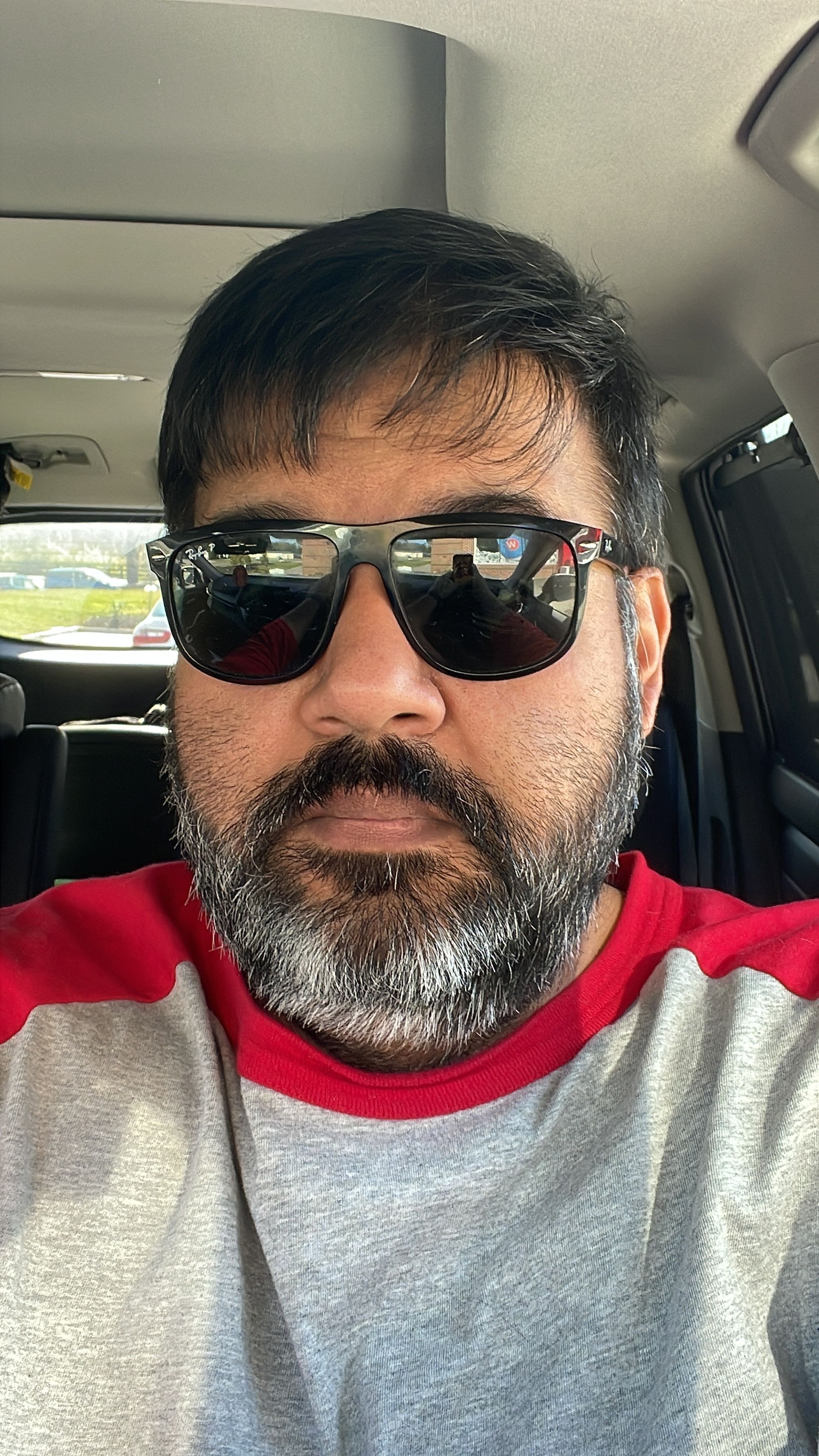 Man with dark hair, beard, sunglasses, and a gray and red shirt, sitting in a vehicle, smiling.